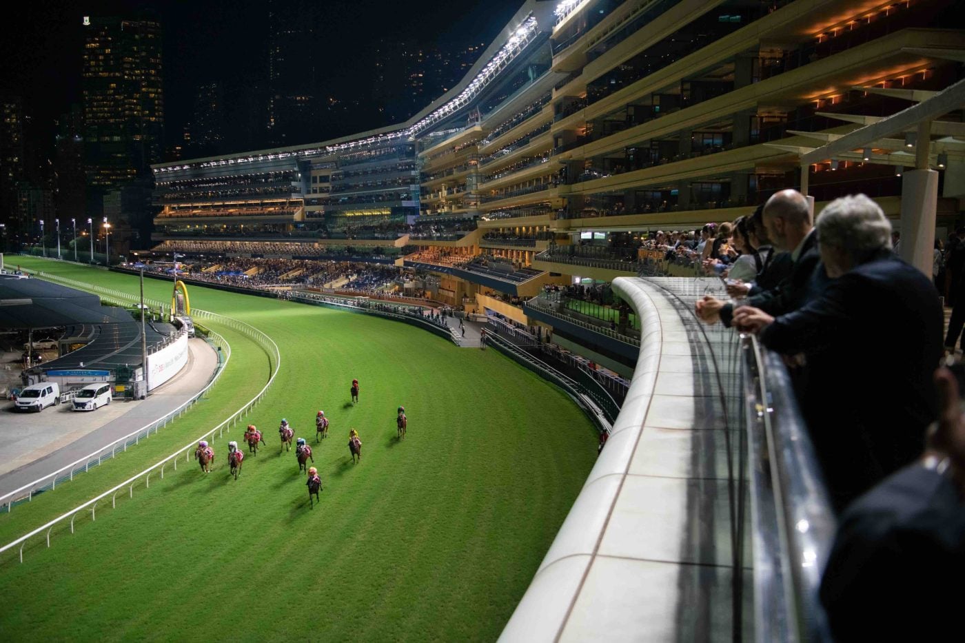 Horses racing under floodlights at Happy Valley Racecourse with city skyline.